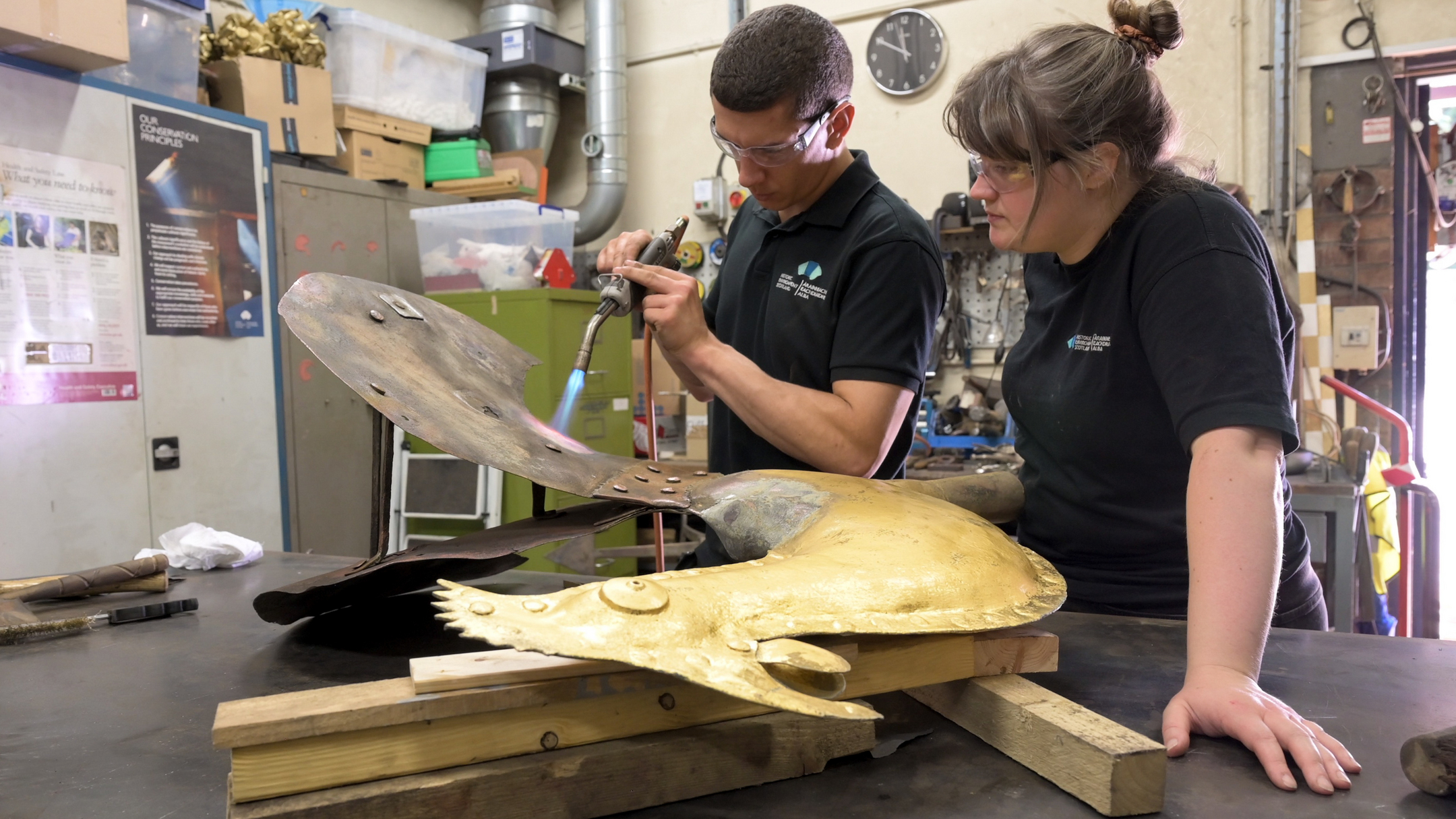 Two conservation staff examining and conserving a large metal weather vane from the top of Glasgow Cathedral, laid out on wooden supports in a workshop with tools and equipment in the background.