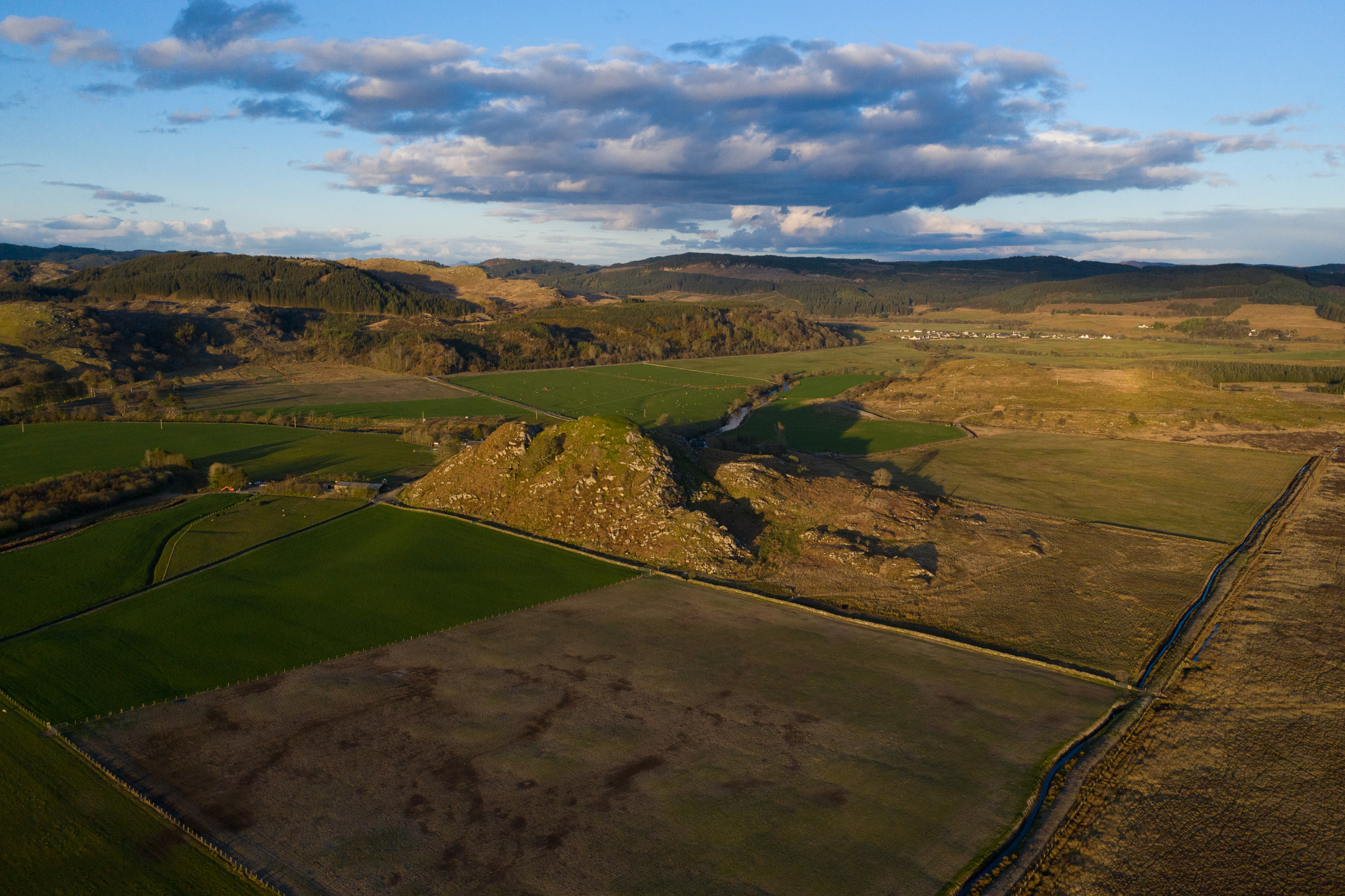 Aerial view of Dunadd Hill Fort on a rocky outcrop, surrounded by a patchwork of green fields, forest, and rolling hills.