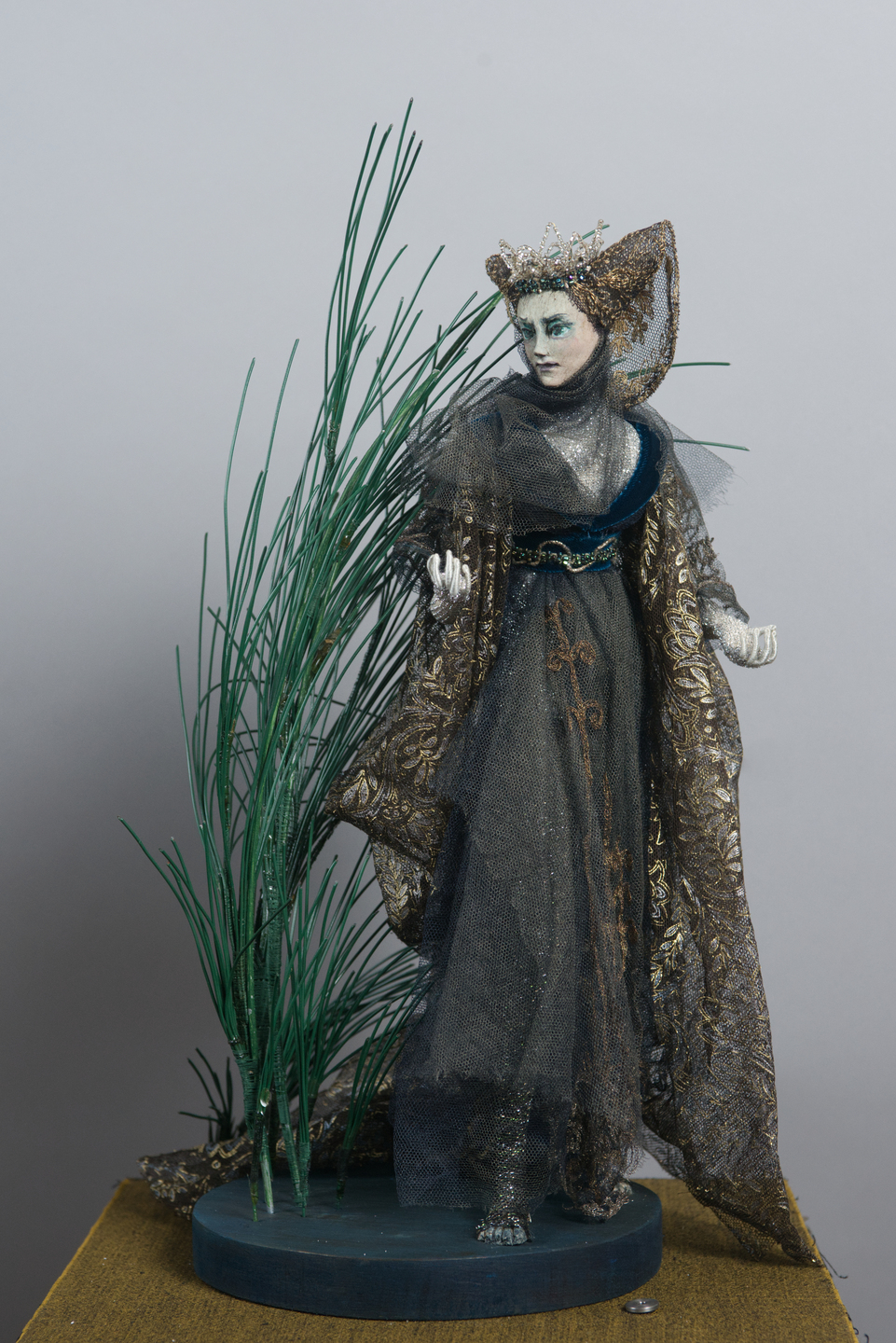 A model dressed in an ornate dark gown with metallic gold patterns and a sweeping cloak, standing beside tall artificial reeds. The figure wears a decorative headdress and appears to be positioned in a dramatic, atmospheric scene.