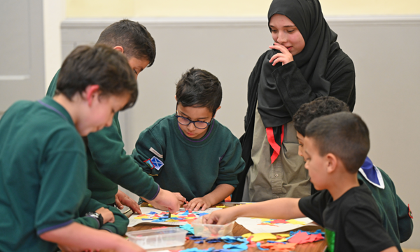 Six kids, some in scout uniforms, sit or stand around a table where they are organising pieces of coloured paper. One is a girl wearing a religious head scarf.