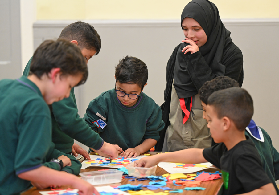 Six kids, some in scout uniforms, sit or stand around a table where they are organising pieces of coloured paper. One is a girl wearing a religious head scarf.