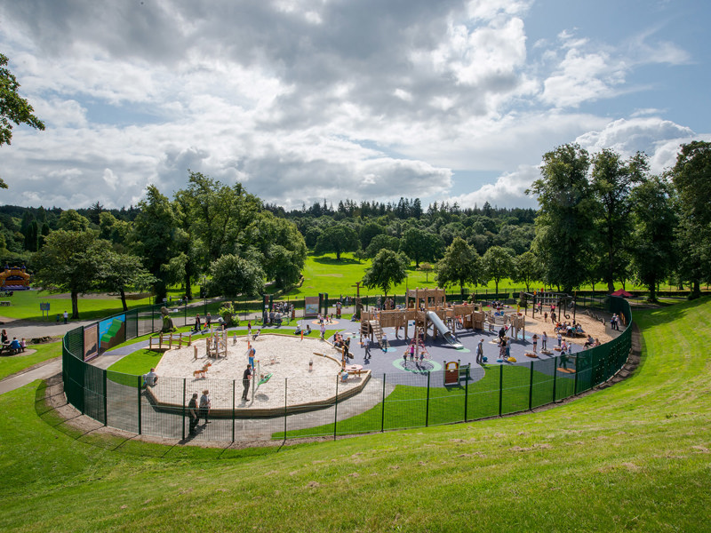 A playground full of children on a sunny day. The playground is surround by a fence and appears to be situated in the middle of a much large park filled with trees.