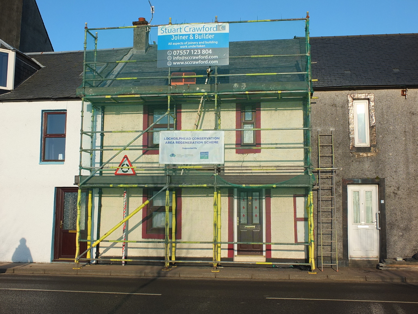 Street‑front view of a two‑storey building covered in scaffolding and safety netting, with contractor signage attached, indicating repair or conservation work underway