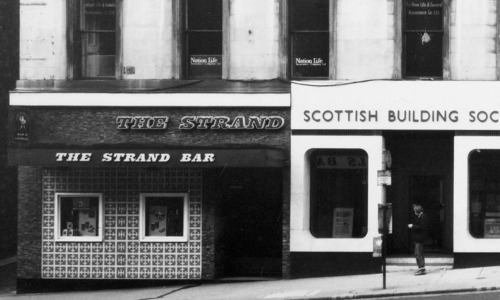 A black and white photo of a four-story stone building with windows. The bottom floor has the words The Strand Bar on awning above the entrance.