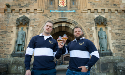 Two men stand outside the front gate of a large stone castle, each wearing a navy and white rugby shirt and holding a small glass of whisky. 