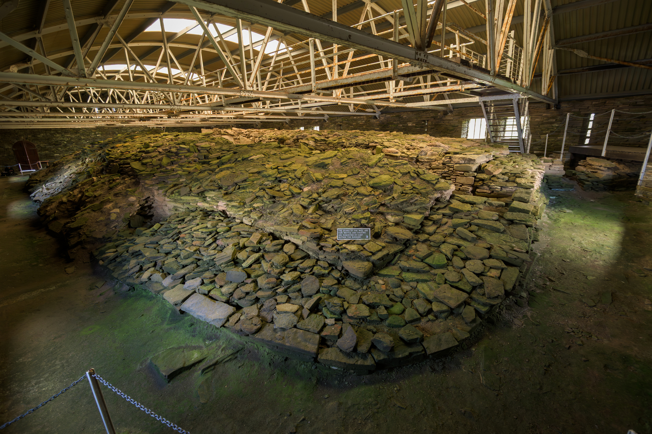 Wide interior view of the cairn showing a mound of stacked stones beneath the roof, with a small interpretation plaque in the centre.