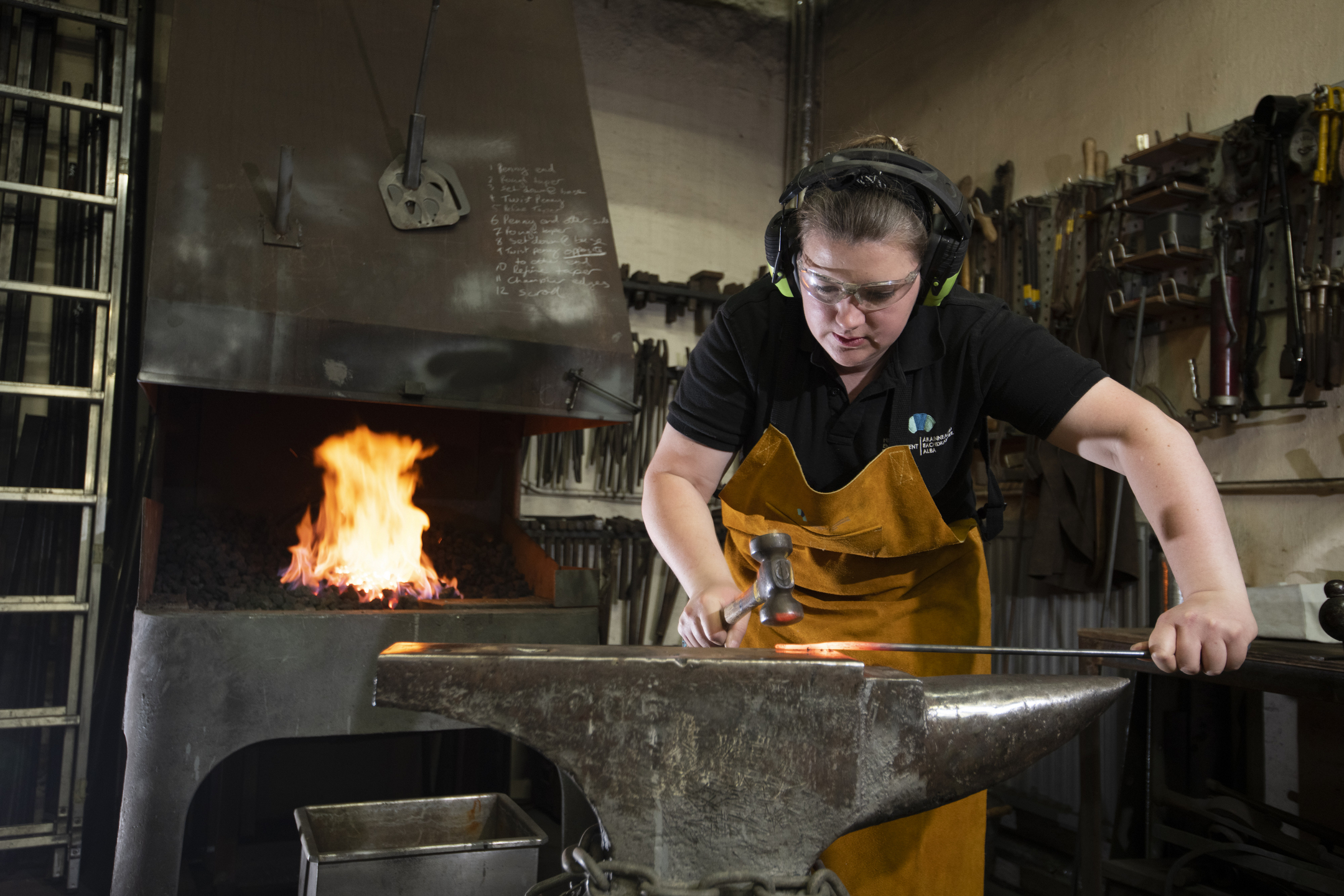 a female blacksmith forging a piece steel on an anvil, a fire is blazing in the furnace behind her.