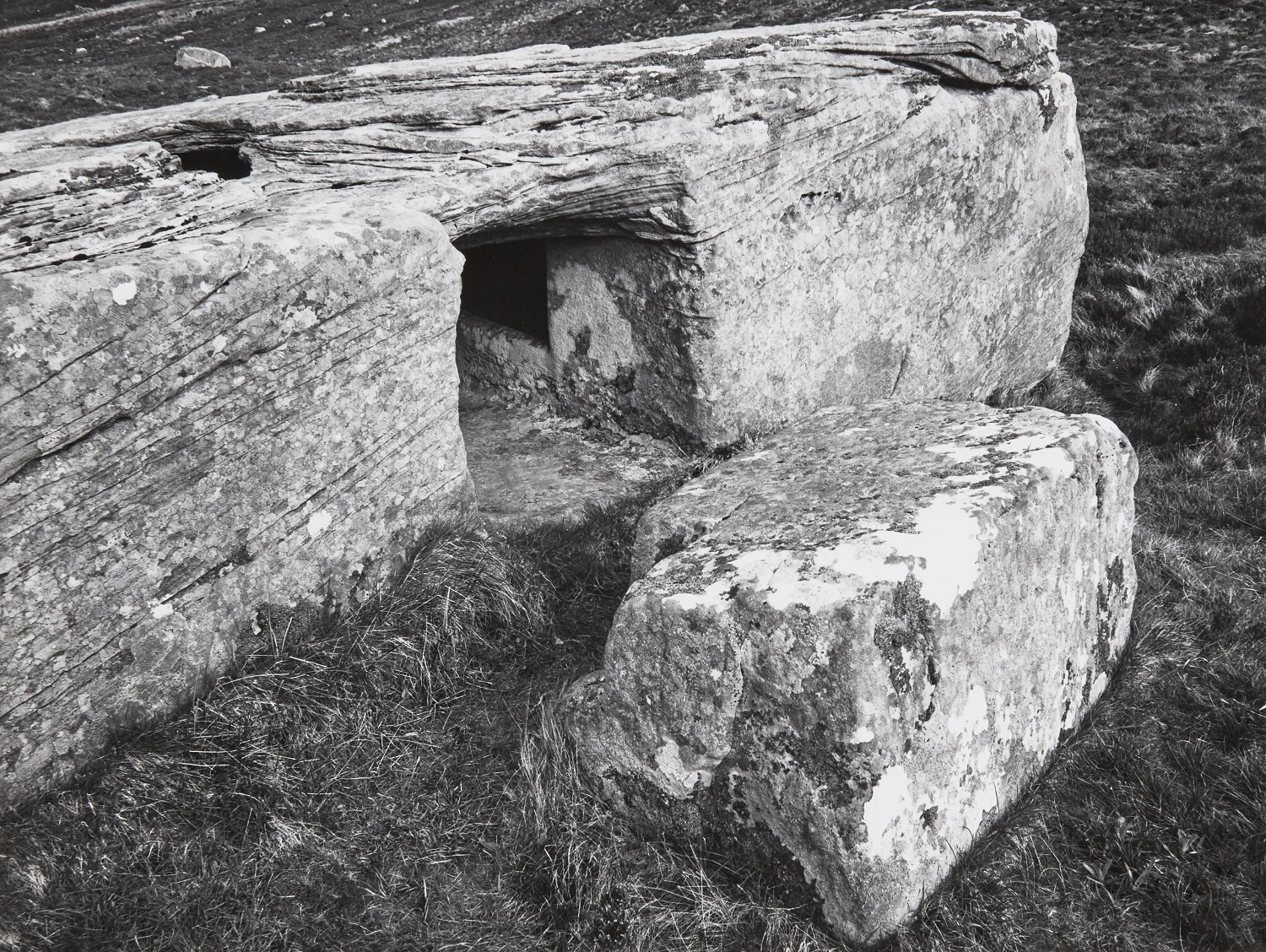 A black‑and‑white photograph showing the low, narrow entrance cut into the side of the Dwarfie Stane, a large rectangular sandstone boulder. A separate block of stone lies in front of the opening, and rough grass surrounds the site.