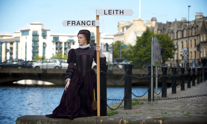 A woman in a historic purple dress holding a sign with two arrows, one pointing to Leith, the other to France