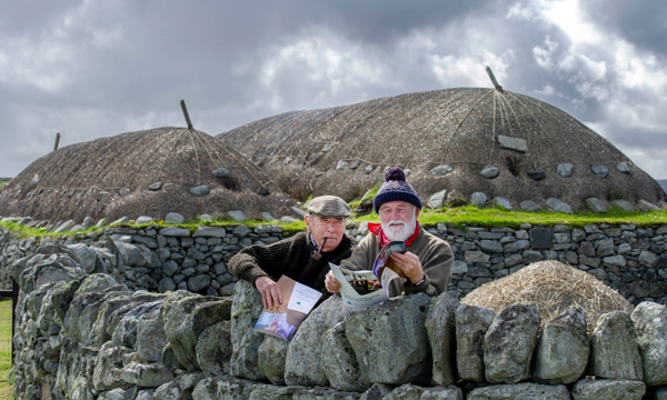 Two men lean on a stone wall in front of Arnol Blackhouse, Isle of Lewis. The man on the left is wearing a flat cap and smoking a pipe. The man on the right is wearing a purple bobble hat and red neckerchief. Both men are holding a copy of Historic Environment Scotland's Gaelic Language Plan.  