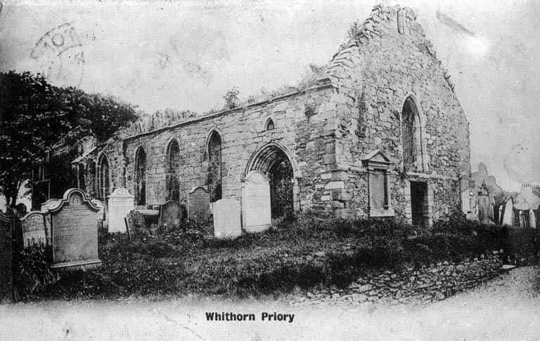 A black‑and‑white photograph of the ruined stone walls of Whithorn Priory, shown alongside an old graveyard with several upright headstones. The structure has tall arched windows and a gabled end with collapsed upper masonry. Printed text below reads “Whithorn Priory.