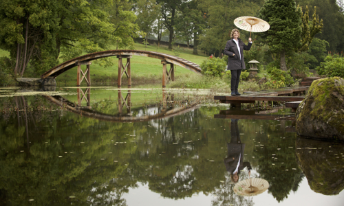 A lady standing beside a lake in a Japenese Garden at Cowden. She is holing a Japenese-style umbrella. A wooden bridge is in the background and its reflection can be seen in the lake. 