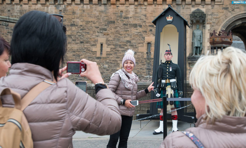 A group of tourists take a mobile photo of a woman in a pink hat, smiling and standing beside a guard wearing a kilt.