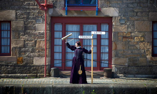 A woman in a historic purple dress stands in front of a building waving a map and holding a sign with two arrows, one pointing to 'France' and one to 'Leith.