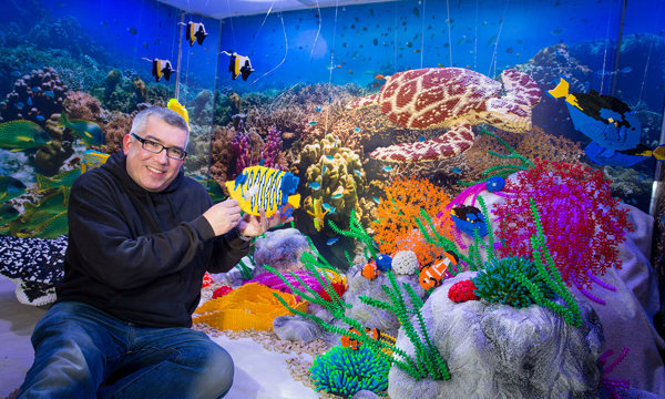 Man sits in front of a large model of the Great Barrier Reef made out of LEGO