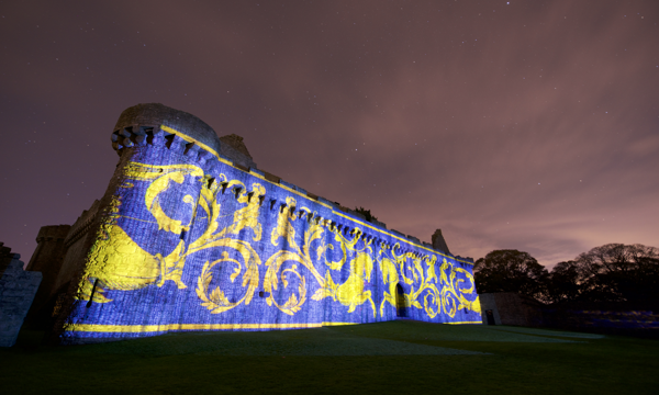 Under a dramatic evening sky, an enormous blue and gold medieval-style pattern is projected onto an exterior wall of Craigmillar Castle