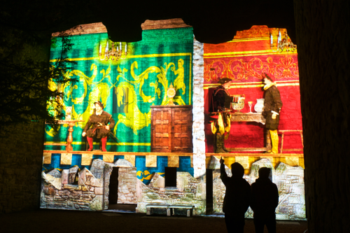 The silhouettes of two people watching a performance of Spotlight on Mary. The action is projected onto a castle wall. On the left hand side, a man with a sword sits at a table in front of a huge green tapestry featuring two gold unicorns. On the right, two men in 16th-century dress converse beneath a chandelier.