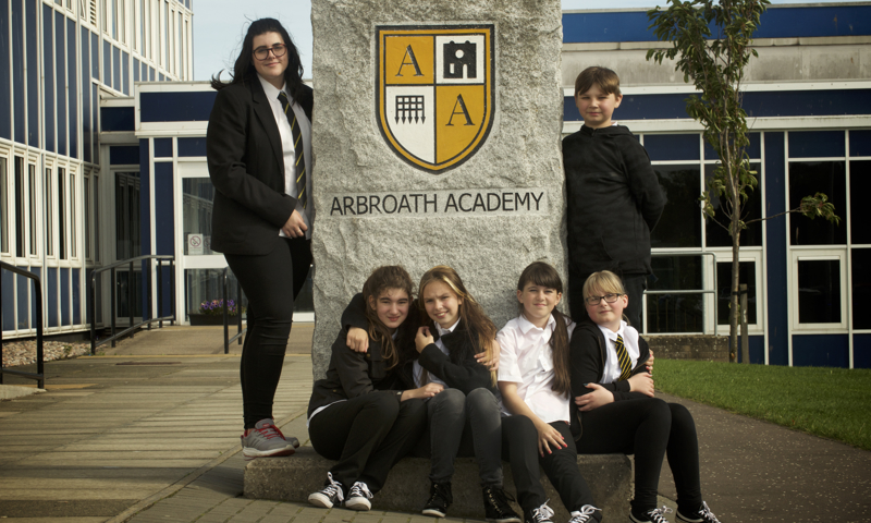 Members of the 1,2 History Crew pose outside Arbroath Academy. They are gathered around a stone sign featuring the Academy