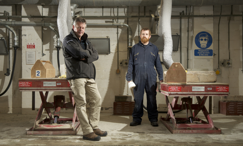 Graham Campbell and Alan Cormie pose inside a workshop. They are each standing beside a red pneumatic lifts upon which sit wooden toolboxes and a piece of masonry. Pipes and ventilation shafts are on the wall in the background along with various warning signs and switches.  