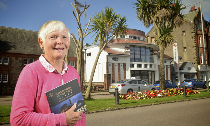 Jane Mayo standing outside Campbeltown Picture House. She is wearing a pink cardigan and holding a copy of a book entitled "Campbeltown Picture House: A Century of Cinema." A colourful flowerbed, some trees and some parked cars can also be seen. 