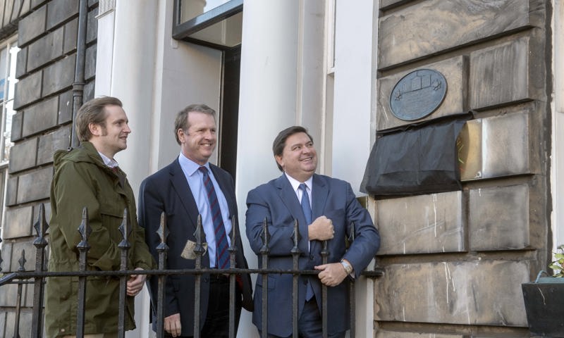 Three men stand on the steps of a Edinburgh townhouse. they have just unveiled a commemorative plaque which is on the wall beside the doorway. 