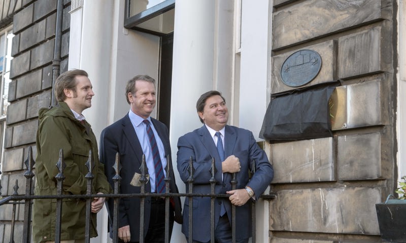 Three men stand on the steps of a Edinburgh townhouse. they have just unveiled a commemorative plaque which is on the wall beside the doorway. 