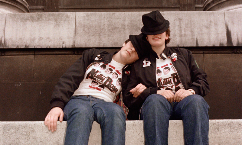 Two teenage girls sitting on the wall of a large building. They are wearing matching outfits consisting of black hats, leather jackets, slogan t-shirts and turned-up jeans. 