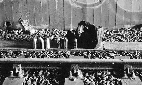 A black and white photo of a rucksack and a line of empty beer cans beside a railway track. 