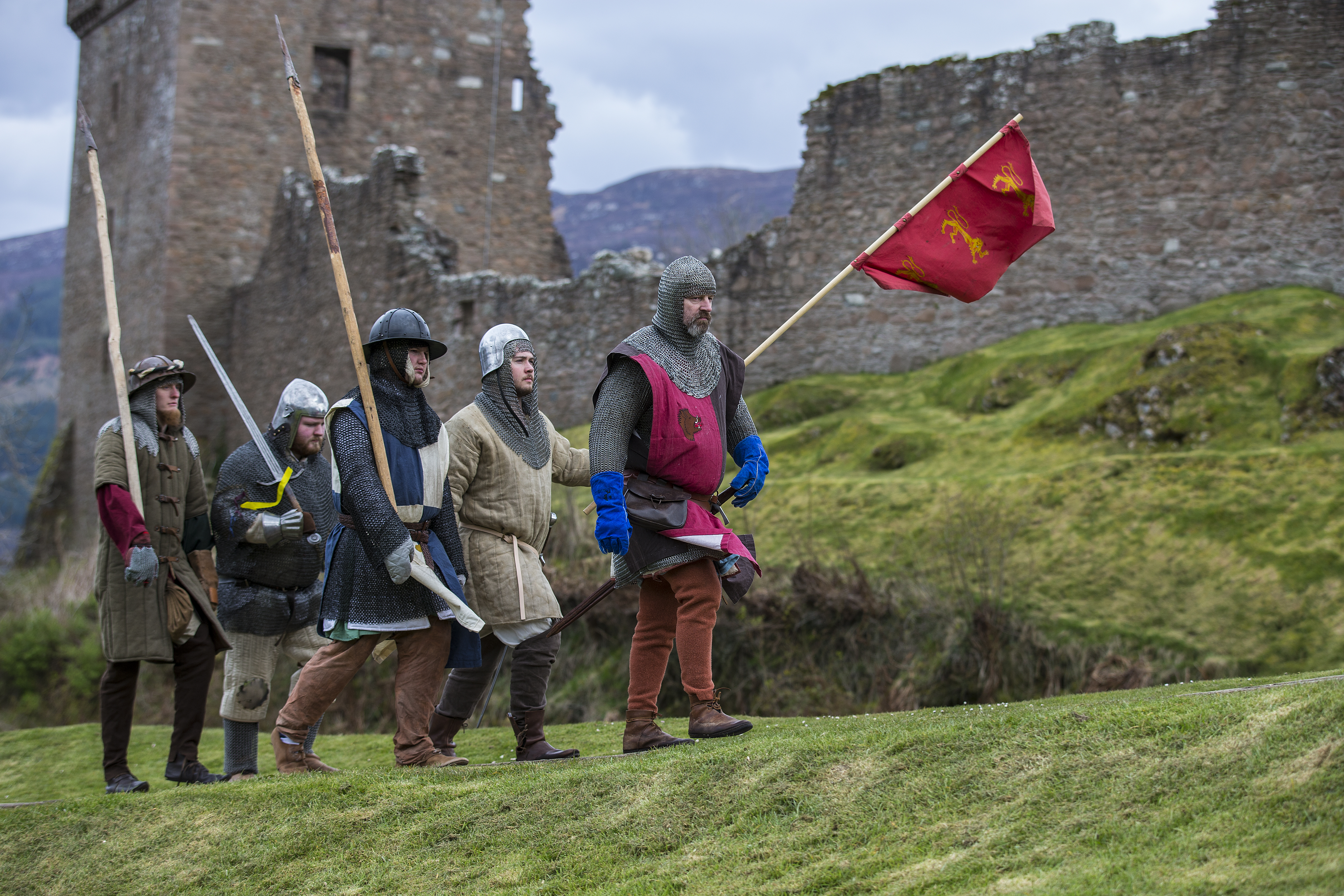 Costumed reenactors performing at an event at a ruined castle. They are wearing medieval military style outfits and hold pikes, swords and flags. 