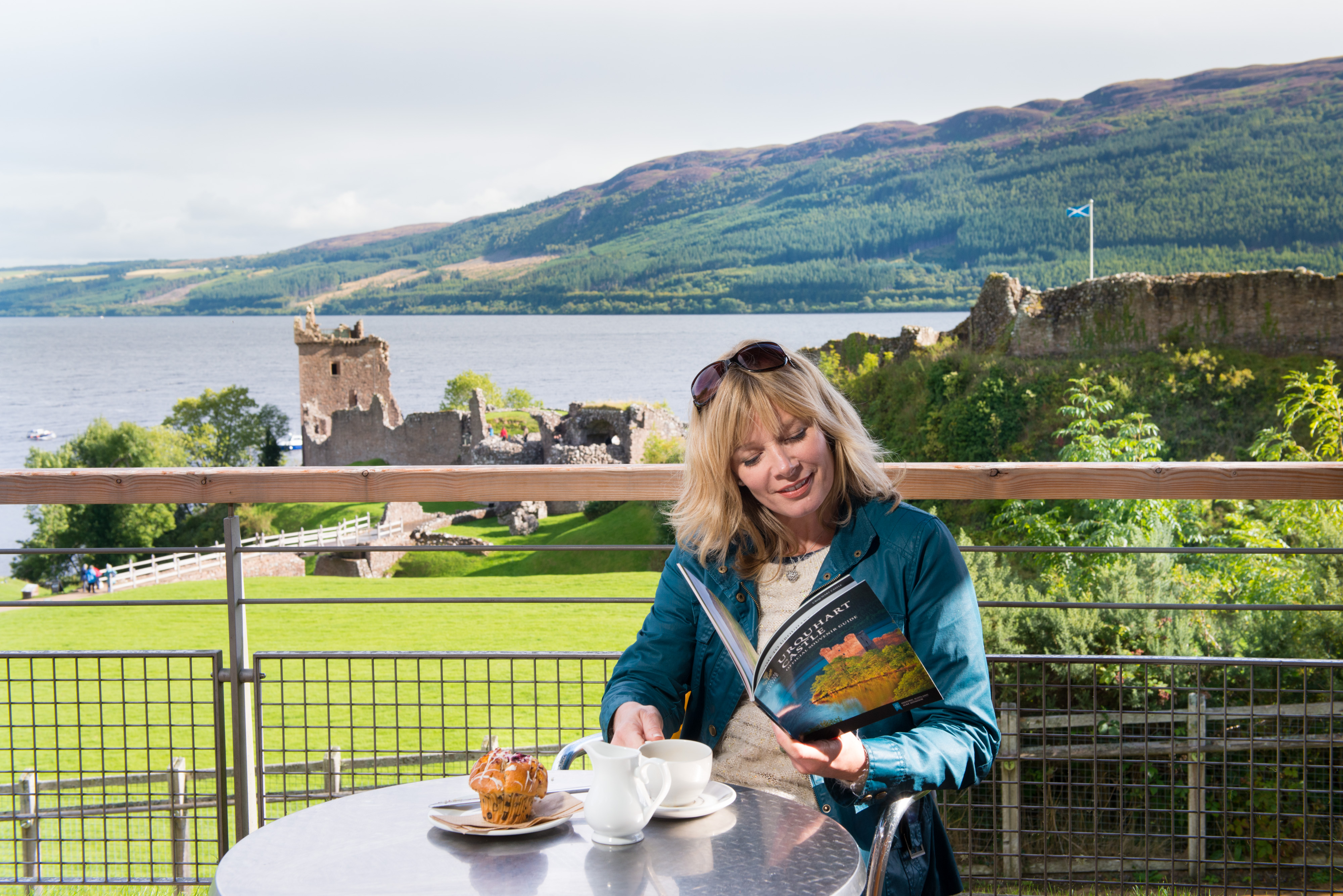 A visitor enjoys a cup of tea at the café at Urquhart Castle. Behind them is a magnificent view of the castle ruins and Loch Ness.