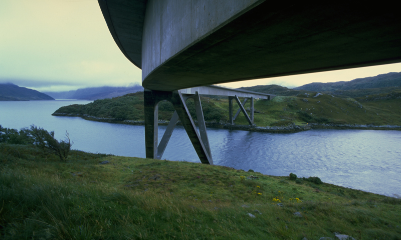 A view of Kylesku Bridge captured from underneath the carriageway. The distinctive curve of the structure can be made out as it crosses the water. 
