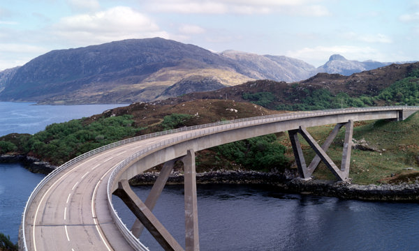A concrete road bridge with a distinctive curve spanning an inlet of water. Mountains and sea can be seen in the background.   