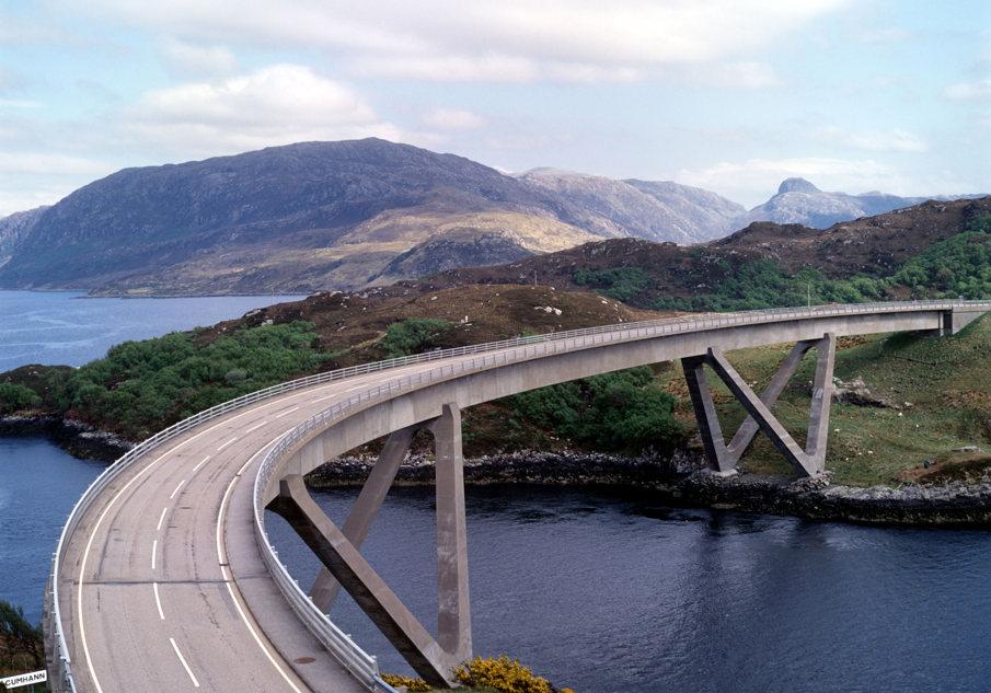 A concrete road bridge with a distinctive curve spanning an inlet of water. Mountains and sea can be seen in the background.   
