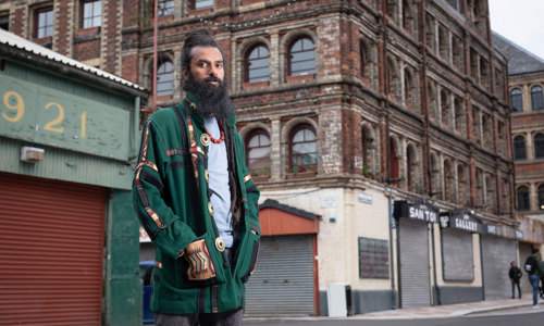 A man with a long beard and dreadlocks stands in front of a red sandstone building.