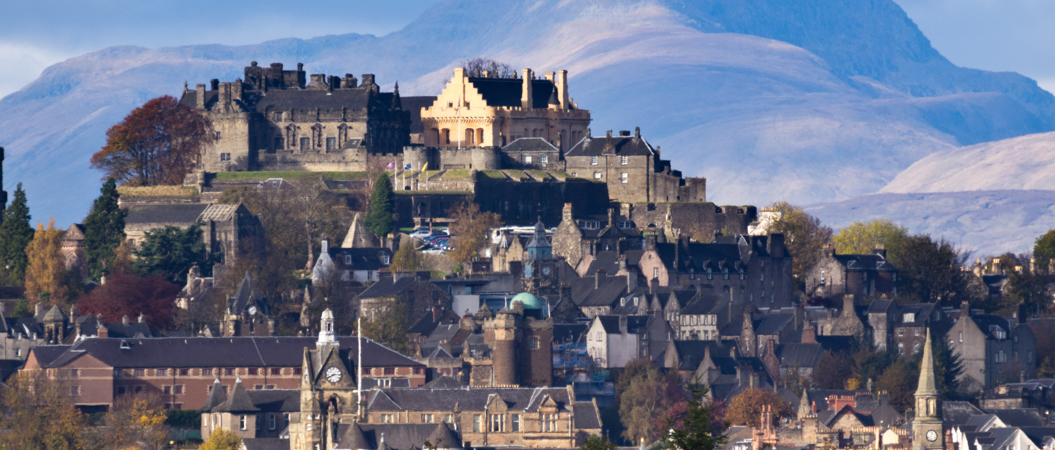 A photo of Stirling Castle towering over the city of Stirling. The distinctive colour of the Castle's Great Hall can be seen. A sunsoaked mountain is in the background.
