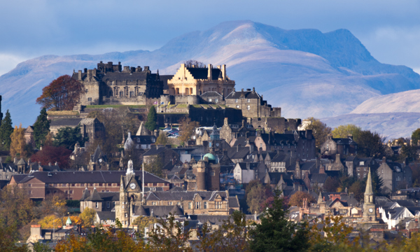 A photo of Stirling Castle towering over the city of Stirling. The distinctive colour of the Castle's Great Hall can be seen. A sunsoaked mountain is in the background. 