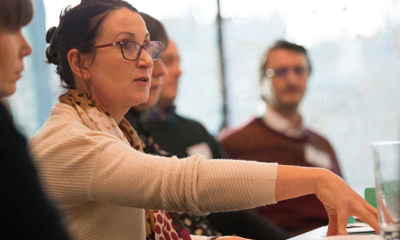 A woman wearing glasses and a cream cardigan speaking at a meeting.