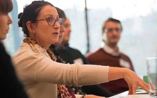 A woman with brown glasses in a beige top speaks to a group of people at a table