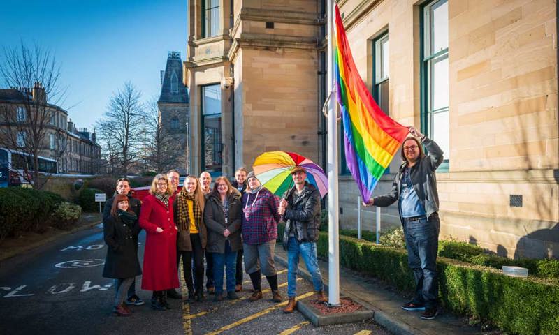 A group of people smiling beside a building and a large rainbow flag.