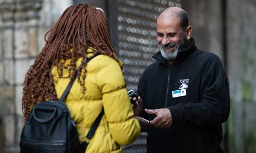 A man with a beard and a name badge smiles at a person in a yellow coat as he scans a ticket.