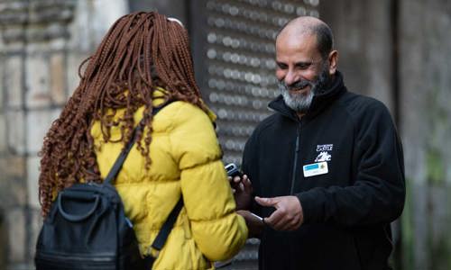 A man with a beard and a name badge smiles at a person in a yellow coat as he scans a ticket.