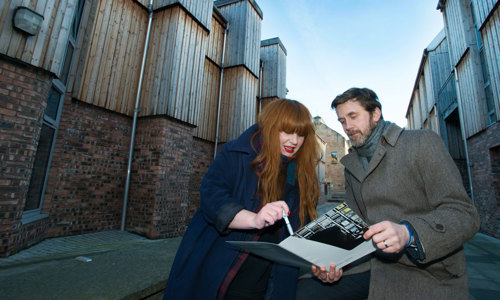 A woman with long red hair and wearing lipstick points at a document held by a man in a brown coat.