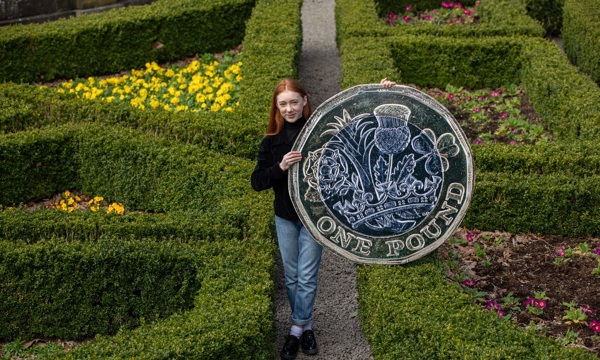 Cherry Campbell, star of Katie Morag, stands in a formal garden holding an oversized £1 coin