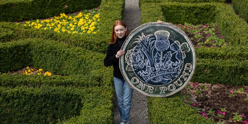 Cherry Campbell, star of Katie Morag, stands in a formal garden holding an oversized £1 coin