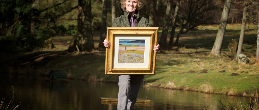 A woman smiles at the camera as she holds a painting of a landscape and stone heart-shaped circle.