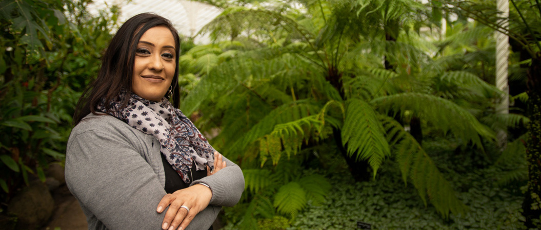 A woman with dark hair and folded arms smiles at the camera. She is standing in a glass building surrounded by greenery.