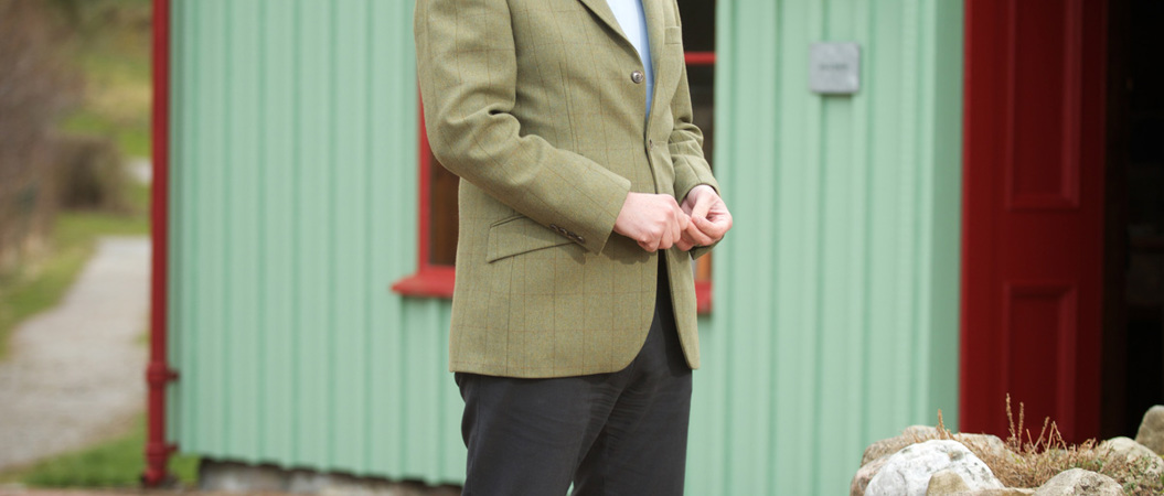 A man in a green jacket is laughing as he stands in front of a green building. There is a sign on the roof which reads "Knockando Woolmills Warehouse"