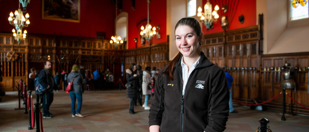 A young woman with long dark hair in a ponytail smiles at the camera. She is wearing a fleece with the words "Edinburgh Castle" written on it and stands in a large hall filled with chandeliers and historic weapons.