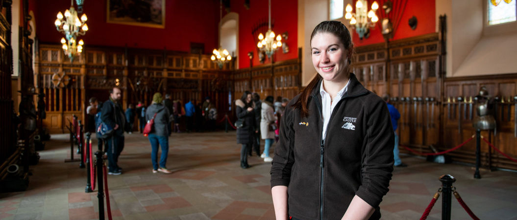 A young woman with long dark hair in a ponytail smiles at the camera. She is wearing a fleece with the words "Edinburgh Castle" written on it and stands in a large hall filled with chandeliers and historic weapons.