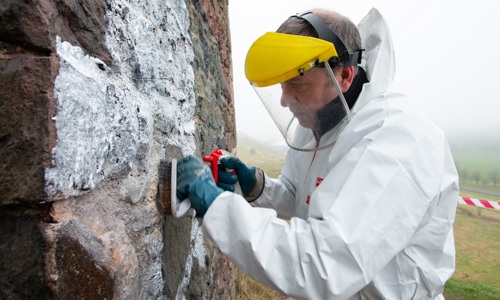 A man wearing a protective visor and bolier suit removes graffiti from a wall